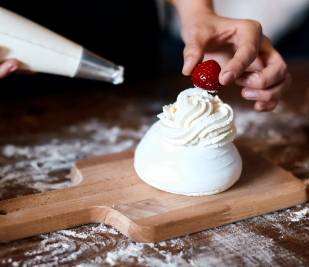 Serving suggestion recipe of a meringue, with Lakeland Dairies Whipped Cream piped on top, with a close up of chef's hand placing a fresh strawberry on top and holding a piping bag of cream in the other hand