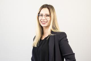 A woman in her 30s with blonde hair smiling wearing glasses, wearing a black jacket and top.