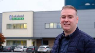 A man smiling in a dark blue shirt and jacket standing in front of a Lakeland Dairies office.