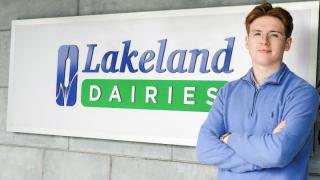 A man in his 20s wearing glasses in front of a Lakeland Dairies sign wearing a blue jumper with his arms folded.